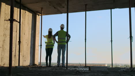 Vista-Posterior-Y-Lateral-Del-Constructor-Caucásico-Con-Casco-Blanco-Y-Chaleco-Verde-Mirando-Un-Dibujo-De-Pie-Sobre-Un-Fondo-De-Construcción-Sin-Terminar.